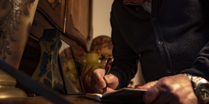 Uomo a mezzo busto che scrive in un quaderno. Ambiente sofisticato con colori tenui.