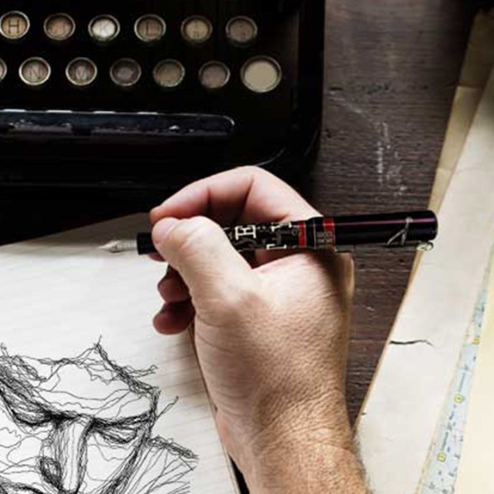 Mano che sta disegnando con penna stilografica Qwerty Visconti davanti ad una vecchia macchina da scrivere.