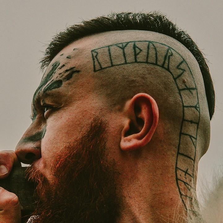 Uomo con caratteri runici tatuati sul lato della testa che soffia dentro un corno