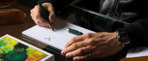 Close up di mani appoggiate su un bloc notes. Una sta scrivendo con una penna stilografica.